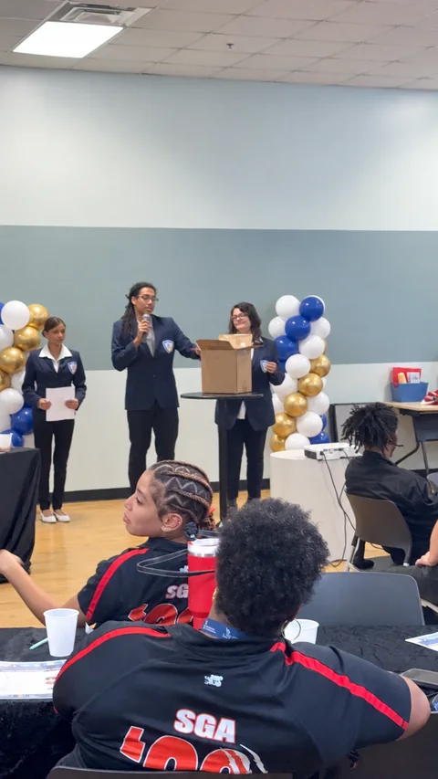 Students in school blazers present a cardboard prototype with a microphone to a seated audience.