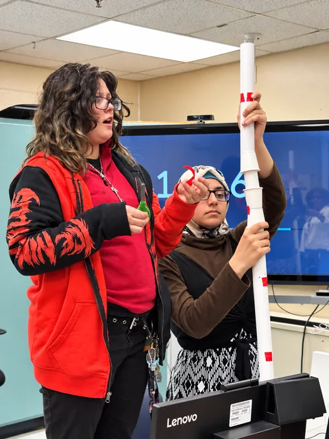 Two students steady a tall paper tower while working with scissors and red tape.