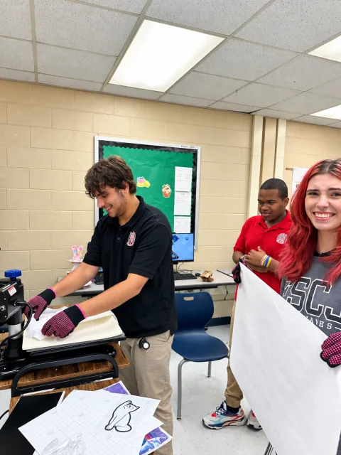 Three students work together at a heat press; one positions a white garment while another holds a large white sheet.